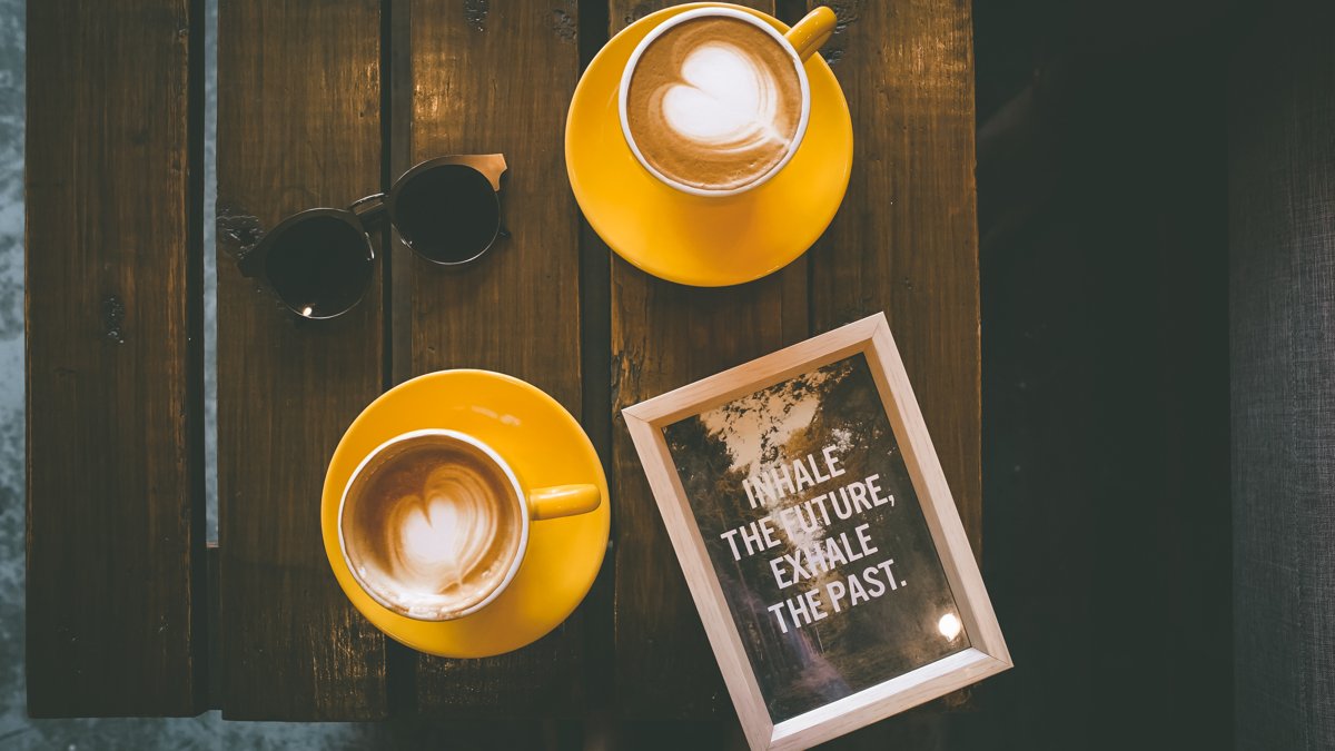Două căni de cafea cu spumă în formă de inimă pe o masă de lemn, lângă niște ochelari de soare și o felicitare înrămată pe care scrie „Inhale the future, exhale the past”