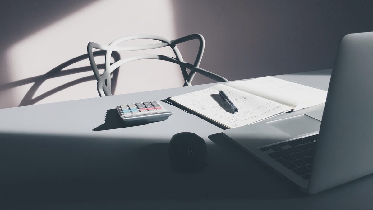 Desk in afternoon light with a calculator, an open notebook with a pen, a mouse and a laptop
